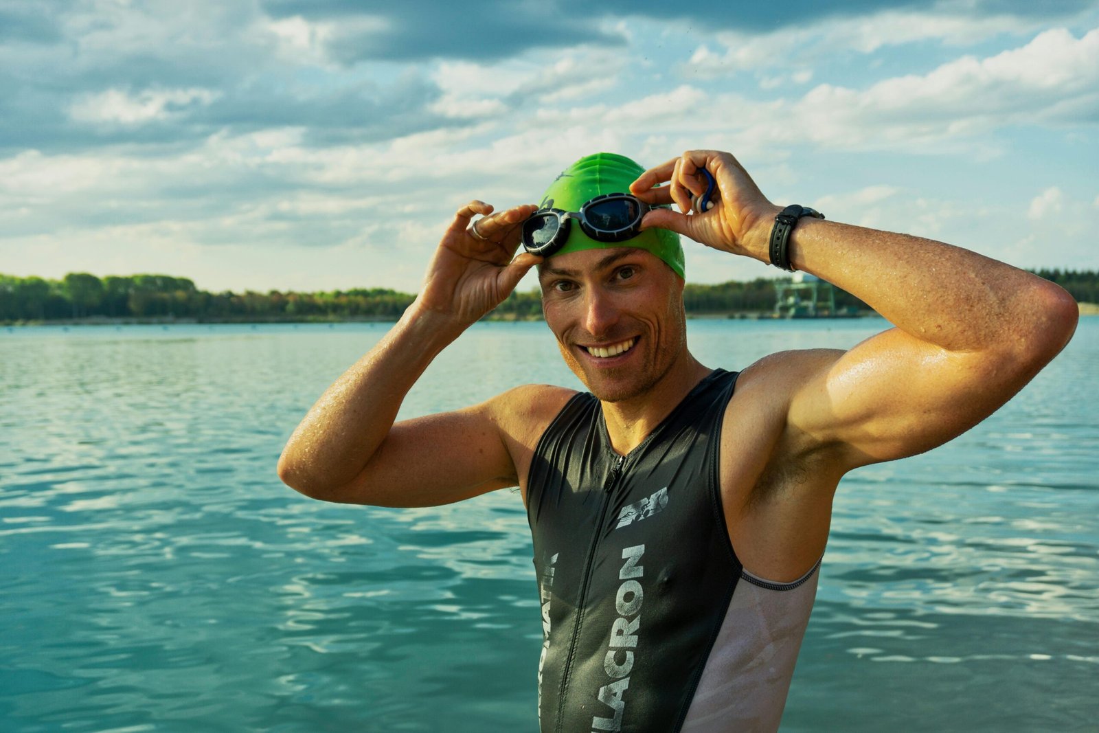 Confident male athlete adjusting goggles at a lake during a sunny day, ready for training.